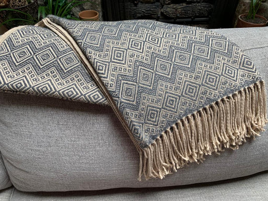 A hand-loomed beige and steel blue alpaca throw blanket with a geometric pattern and fringe, draped over a couch.